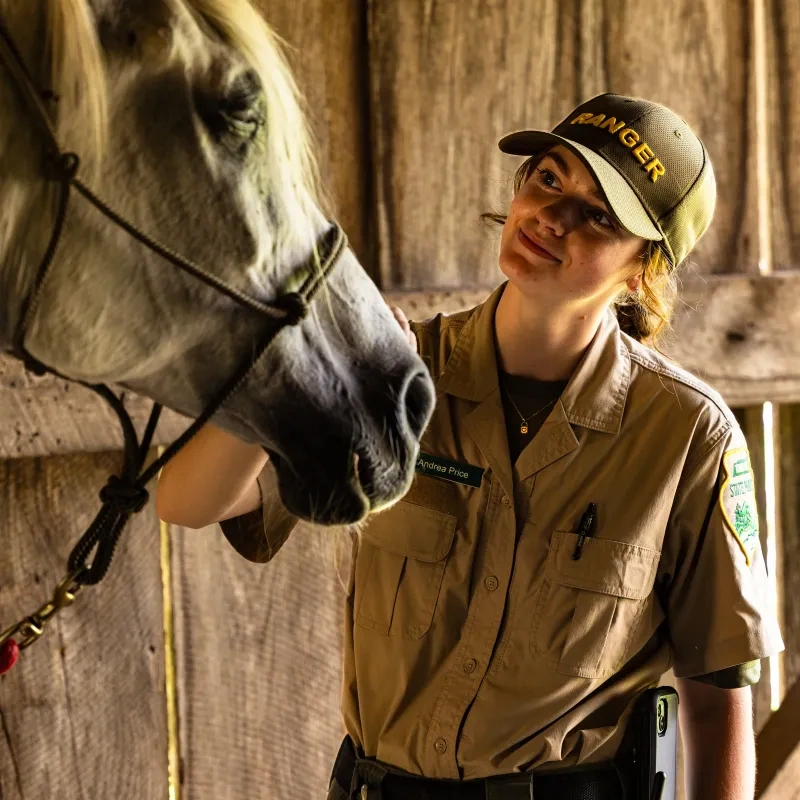 Ranger in uniform petting a white horse inside a wooden barn.
