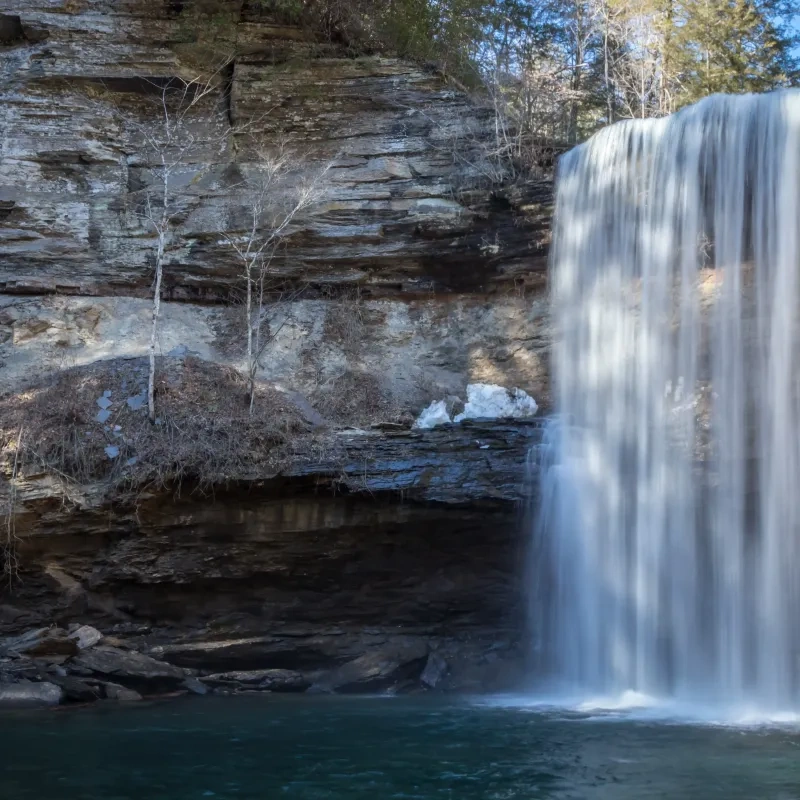 A serene waterfall cascades over a rocky cliff into a calm pool below. Sunlight illuminates the cliff, highlighting the texture of the rocks and creating a peaceful atmosphere.