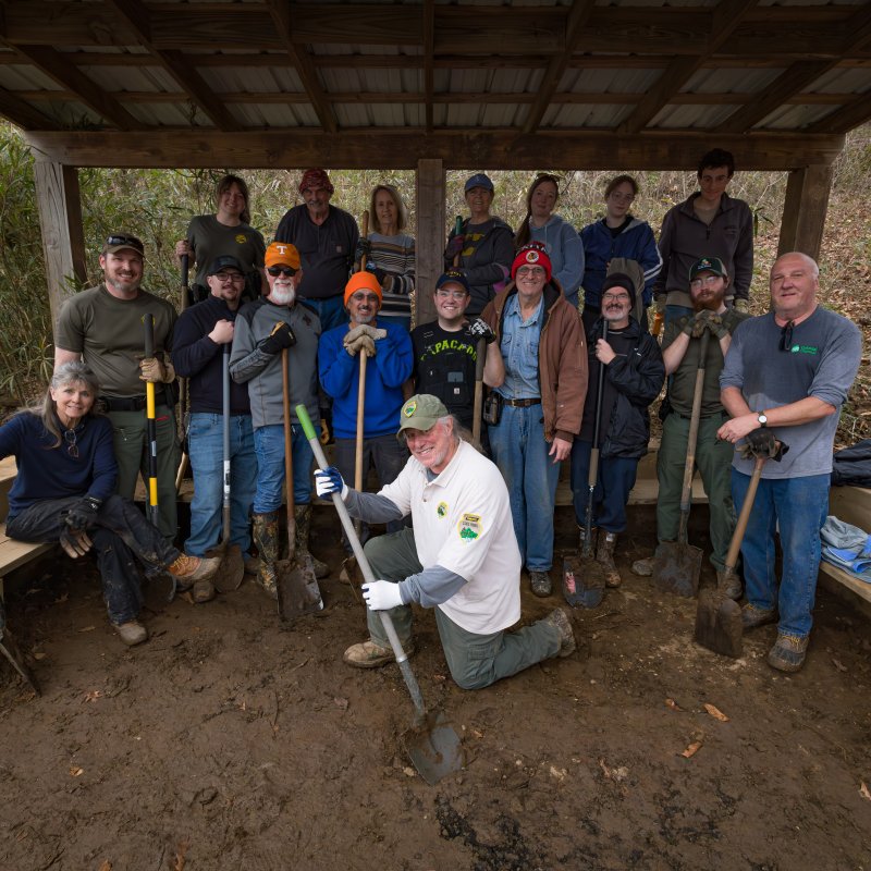 A group of 16 people, smiling and holding tools, poses under a wooden shelter in a wooded area. The mood is upbeat and collaborative.