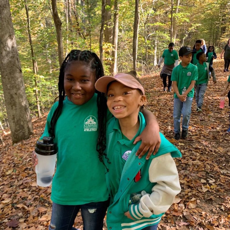 Young girls hugging each other in the forest in front of other kids wearing green in a forest
