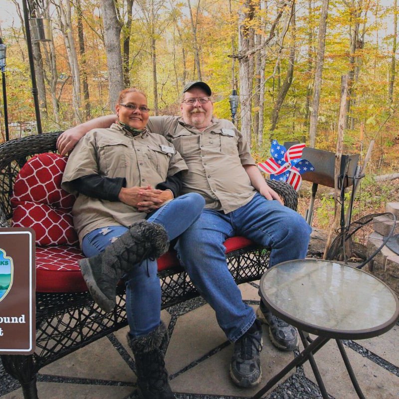 Two people relax on a wicker chair, enjoying the warmth of a fire pit in a cozy outdoor setting.