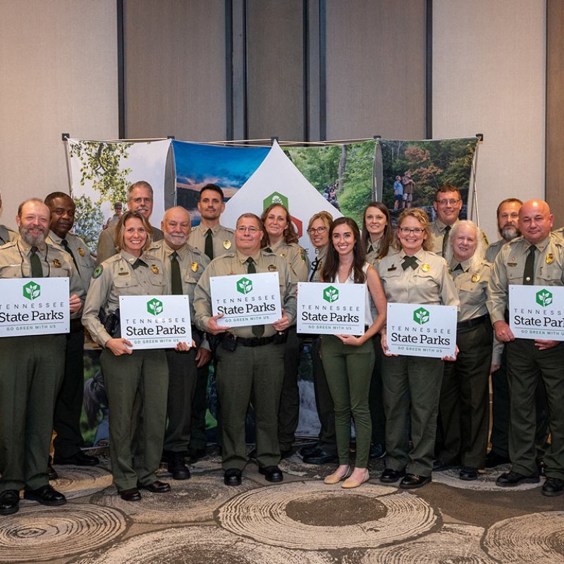 A group of people in uniform stand smiling, holding "Tennessee State Parks" signs. The setting is a decorated indoor event space, conveying a celebratory tone.