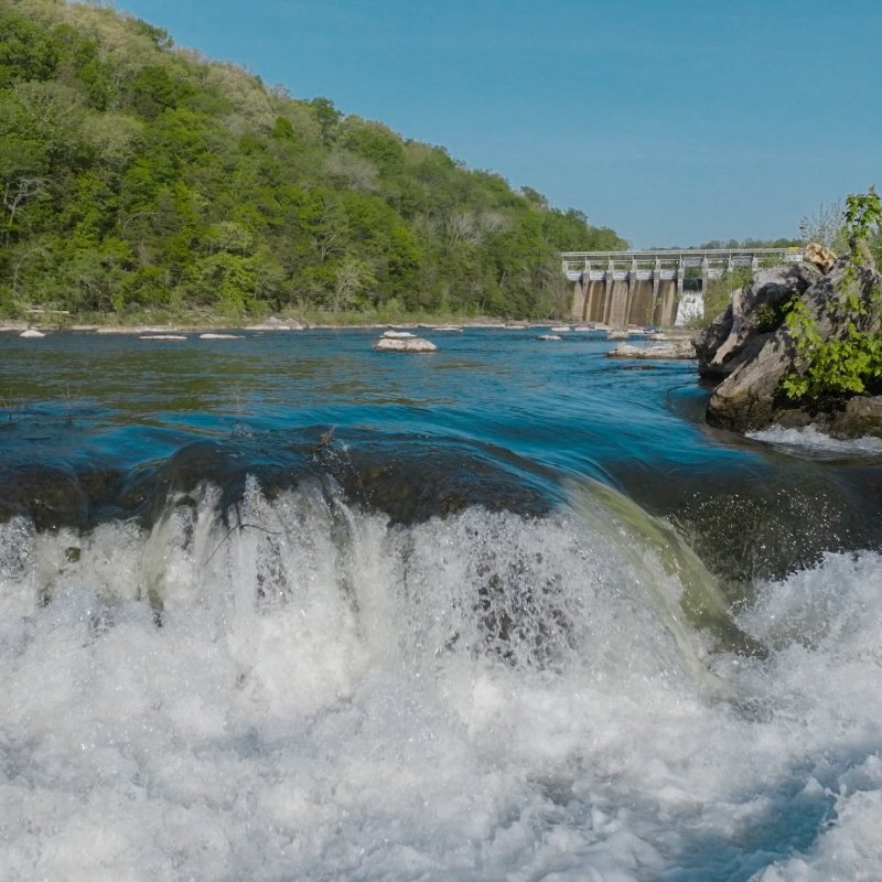 A scenic river with white cascading rapids flows toward a dam. Lush green trees line the banks under a clear blue sky, conveying a serene atmosphere.