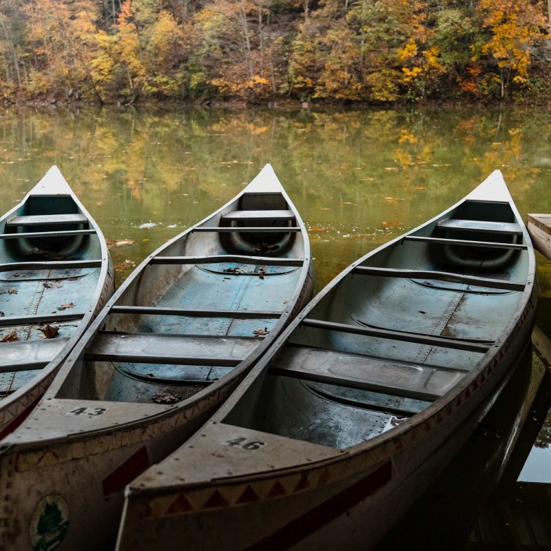 Three canoes are docked under a wooden roof alongside a calm lake, reflecting autumn trees with orange and yellow leaves. The scene feels peaceful and serene.