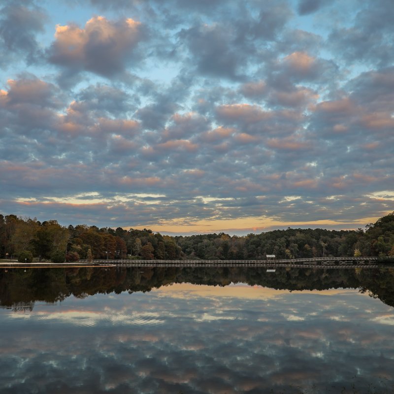 Calm lake reflecting a cloudy, colorful sky during sunset. Surrounding trees in autumn hues line the shore, creating a peaceful, serene scene.