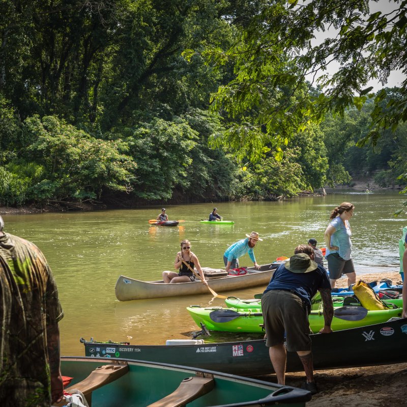 People launch canoes and kayaks into a calm river surrounded by lush green trees, creating a lively and adventurous atmosphere.