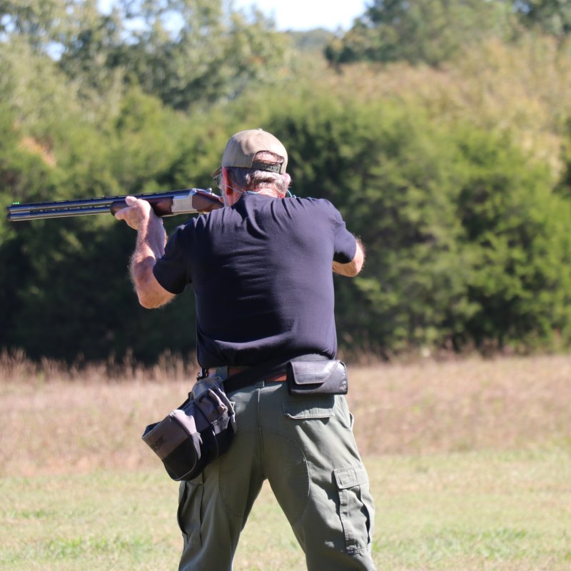 A person standing in a grassy field, aiming a shotgun into the distance. They're wearing a cap and tactical gear, with trees in the background.