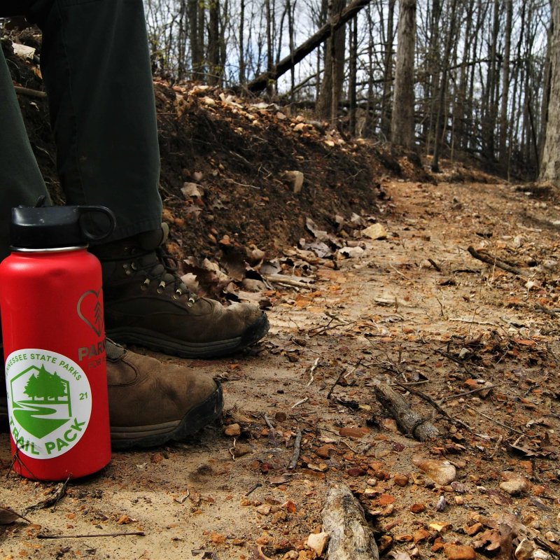 A person wearing hiking boots and green pants sits on a wooded trail. A red water bottle with a nature design is placed beside them, surrounded by fallen leaves.