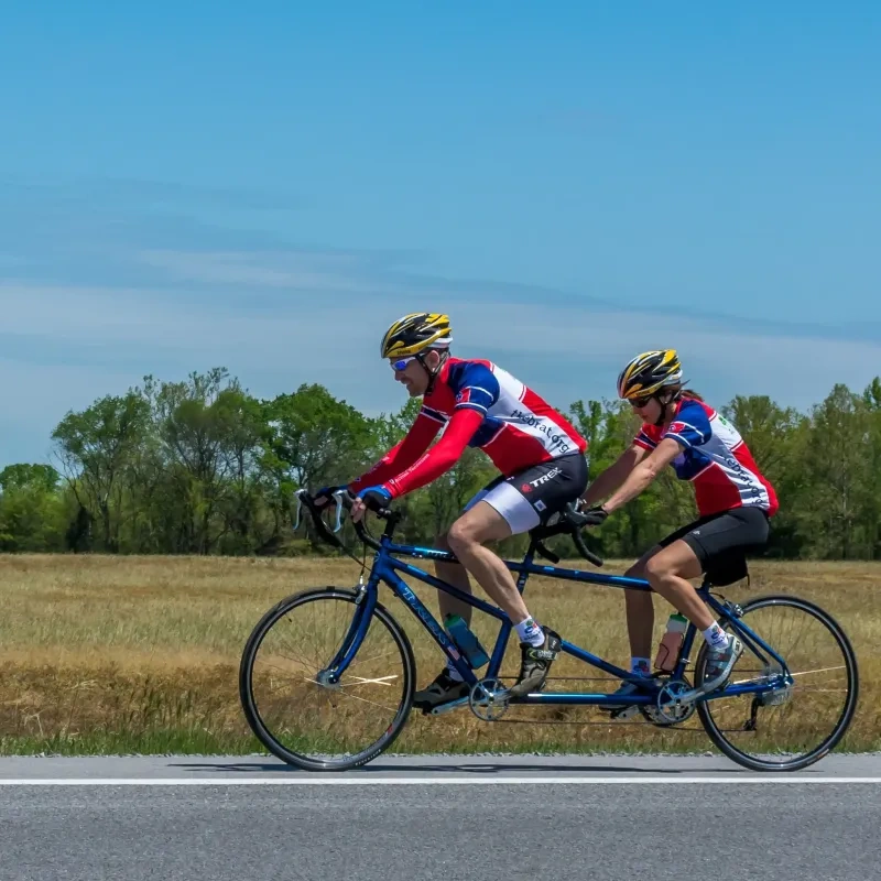 Two cyclists on a tandem bike in a rural area under a clear blue sky.