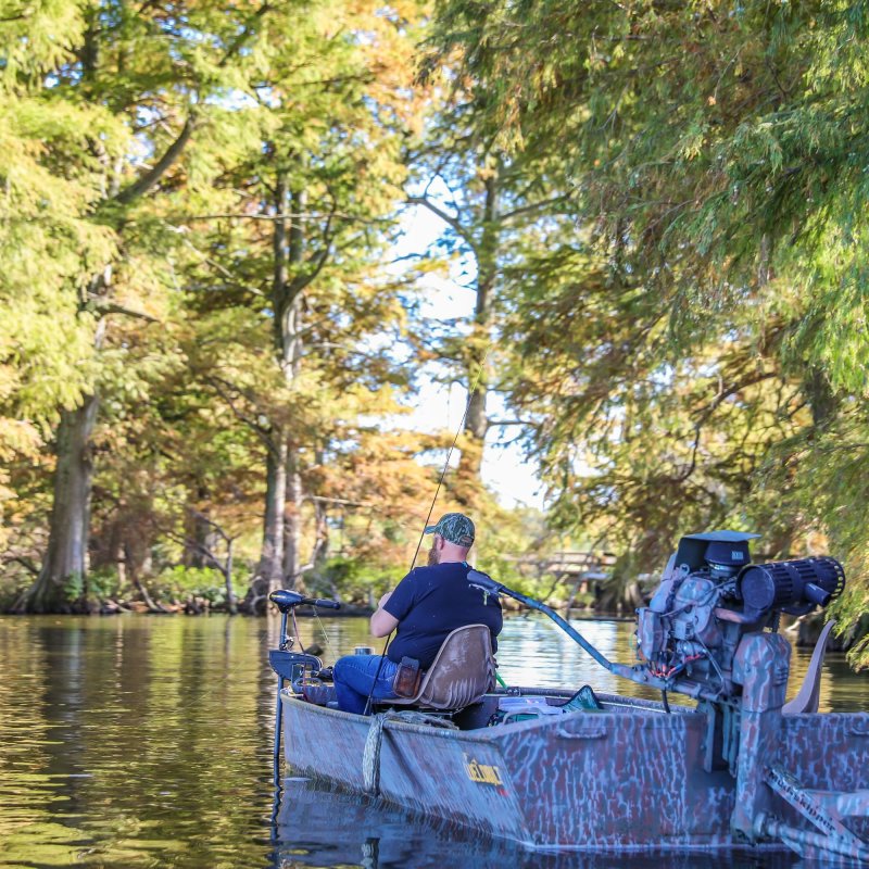 A man in a blue shirt sits in a small motorboat, fishing on a calm river surrounded by tall, lush trees tinged with autumn colors, evoking a peaceful, serene atmosphere.