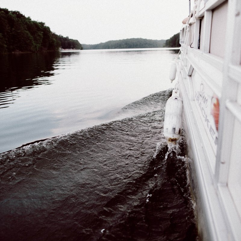 View from the side of a boat cruising on a calm lake, with gentle ripples trailing behind. Lush green trees line the distant shore under a gray sky.