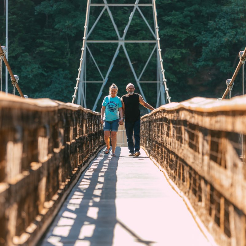 man and woman walking on a narrow bridge towards the camera with trees in the background