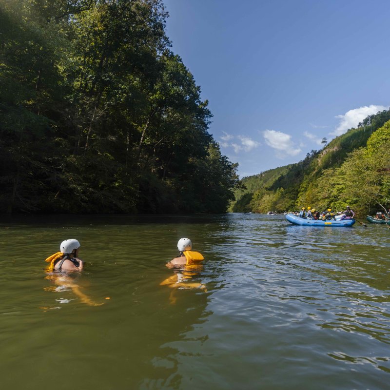 two people swimming in the water watching people in rafts drift off in the distance