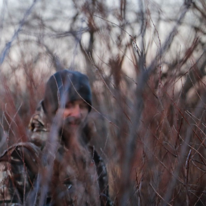 A person in camouflage, partially obscured by dense, bare branches, suggesting a winter setting. The atmosphere is quiet and secluded.