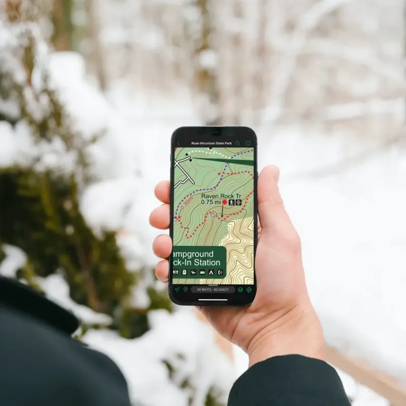 A person standing in a snowy forest holding a smart phone displaying an offline trail map 