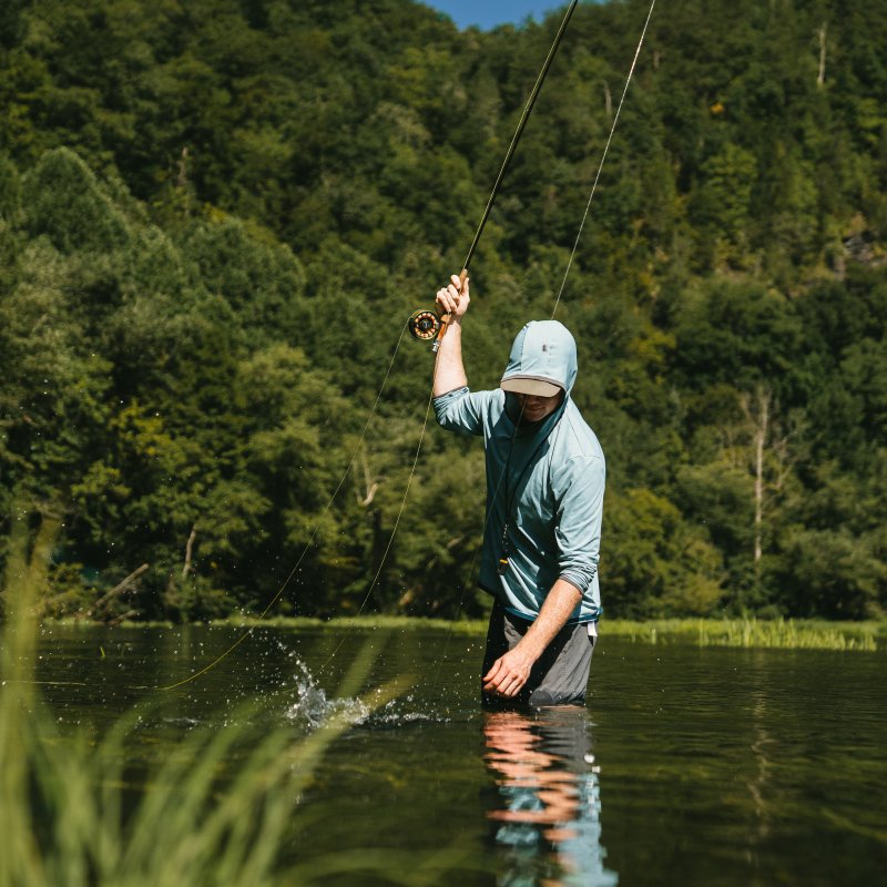 Man fly fishing in a river, surrounded by lush green trees.