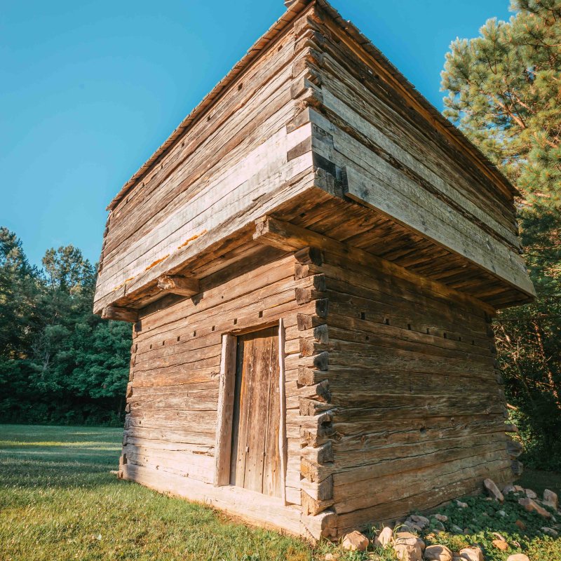 Wooden cabin with skewed upper story in a lush green field.