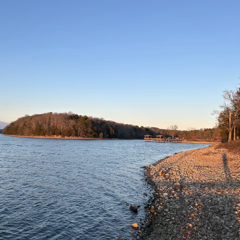 Lakeside view at sunset with rocky shoreline and distant forest.