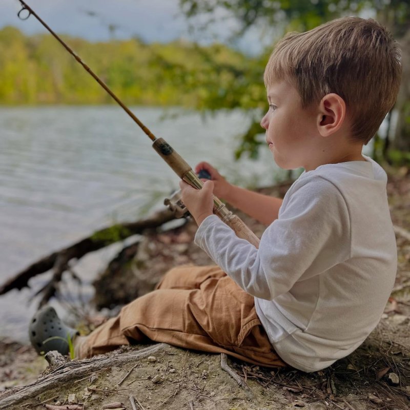Young boy fishing by a lake, seated on a log.