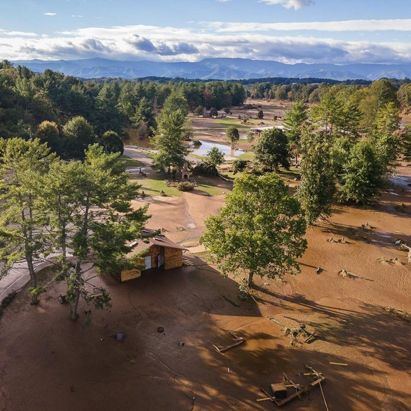 Aerial view of a dry riverbed with sparse trees and a small cabin, mountains in the background.