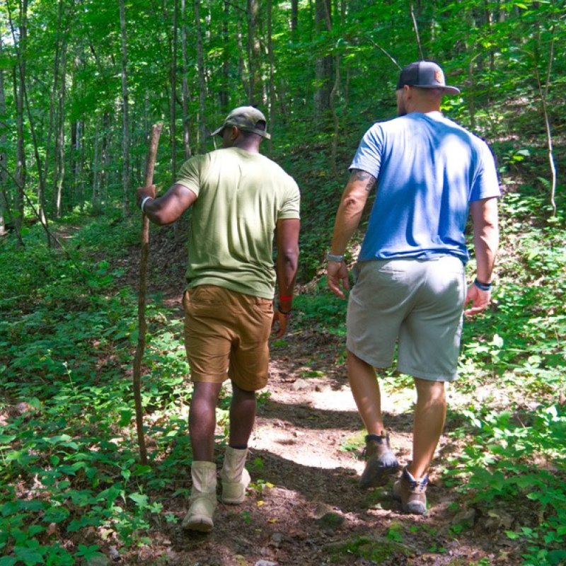 Two men walking along a forest trail, one holding a walking stick.