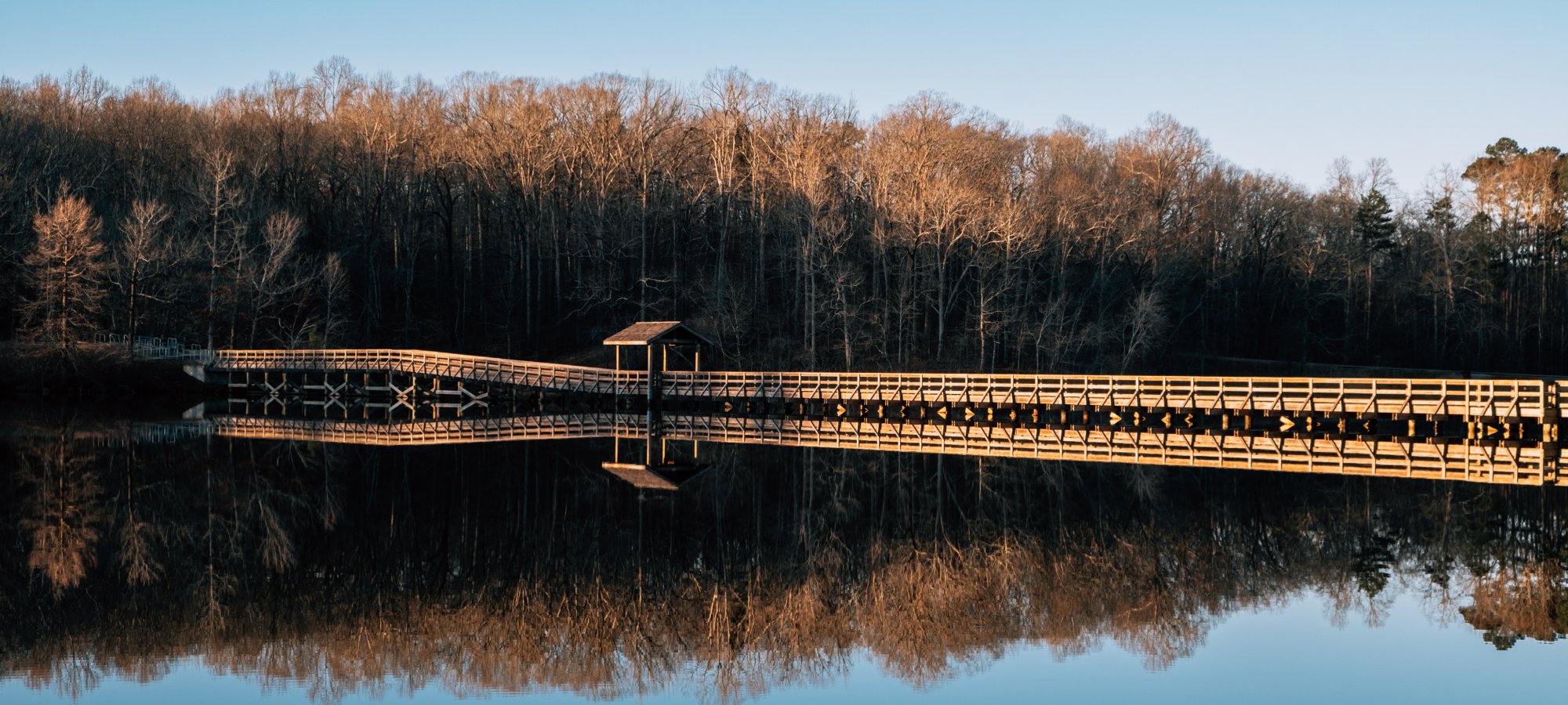 A wooden pier extends over a calm lake with a small pavilion and is mirrored in the water. Bare trees with golden light line the shore under a clear blue sky.