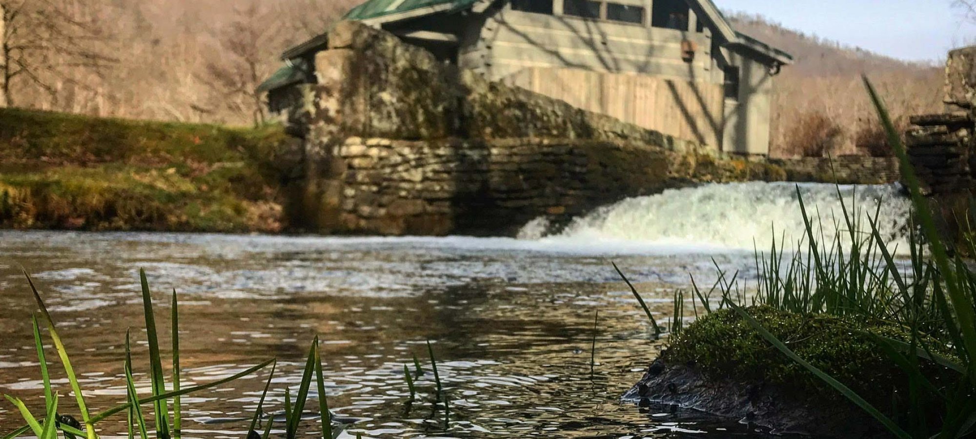 point of view from the river by the mill house where there is a man made waterfall and a stone wall barrier