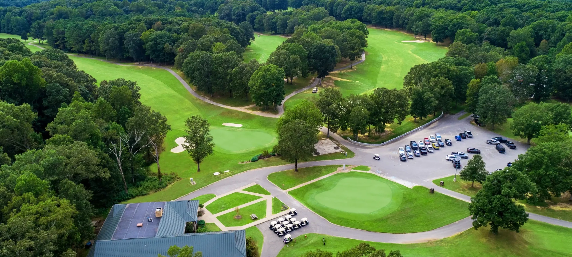 Golf course with greens, clubhouse, parking lot, and surrounding trees.
