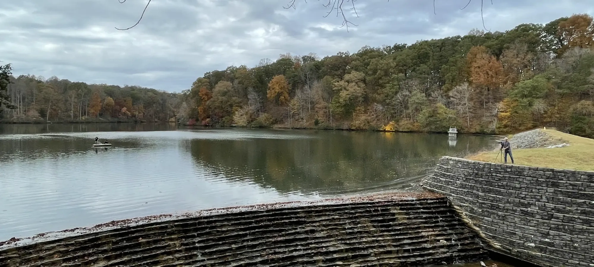 Overcast lake with colorful autumn trees and a dam; a person with a tripod on the right.