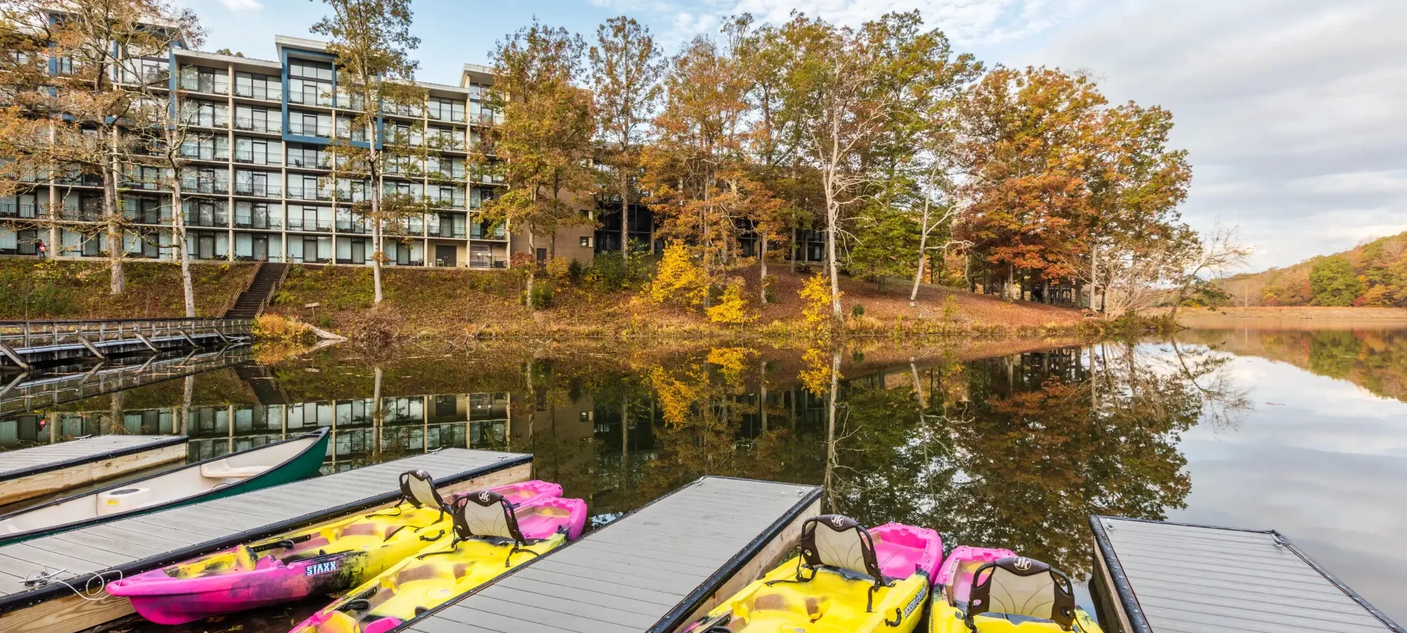Kayaks docked by a calm lake, with a modern building and autumn trees in the background.