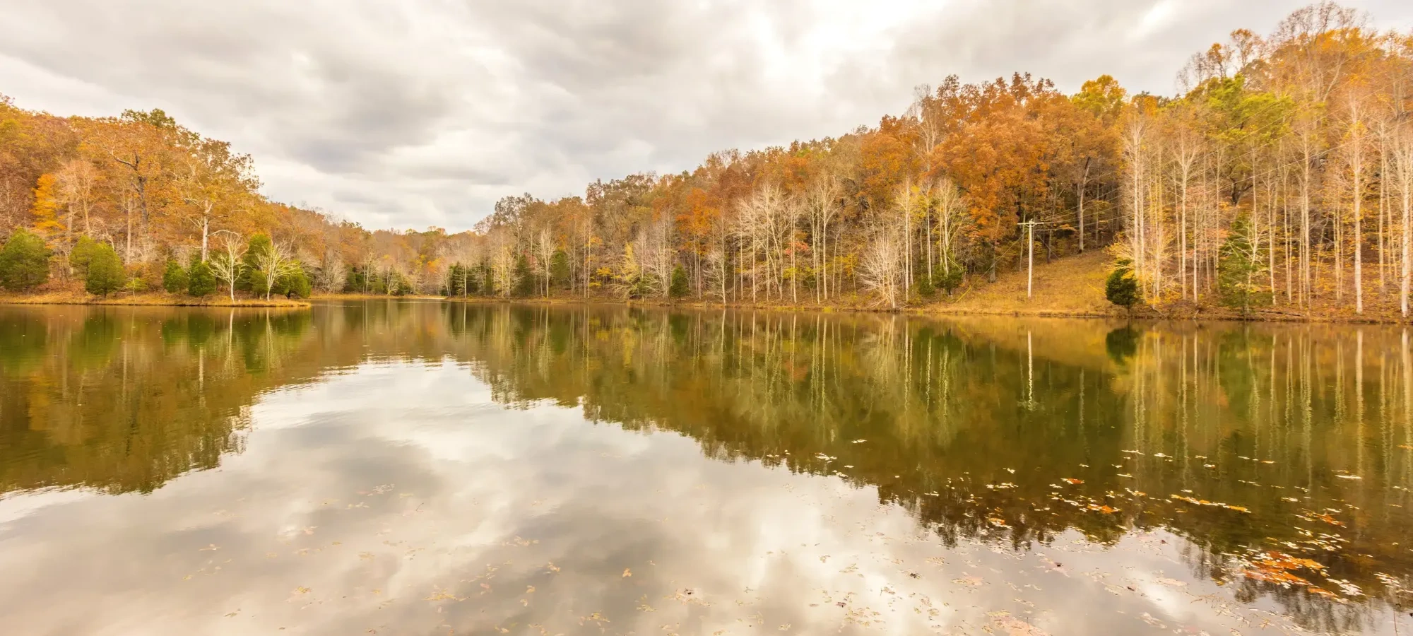 Calm lake reflecting autumn trees and cloudy sky.