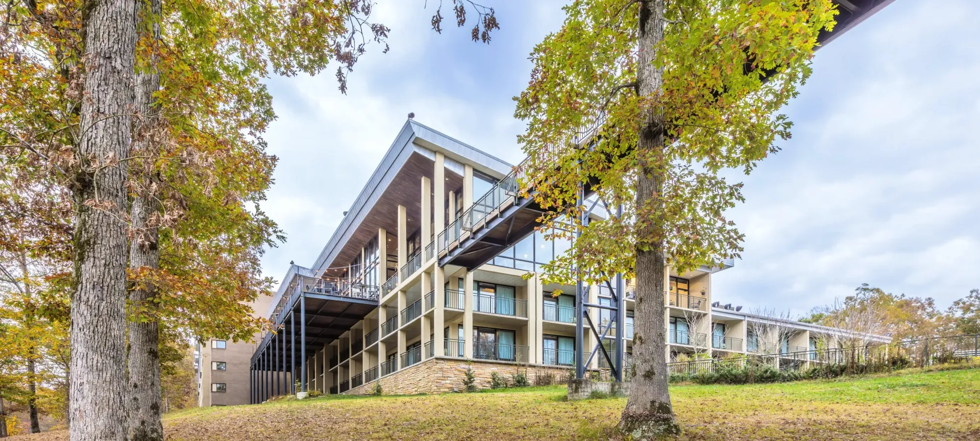 Modern building with large windows, surrounded by autumn trees and cloudy sky.