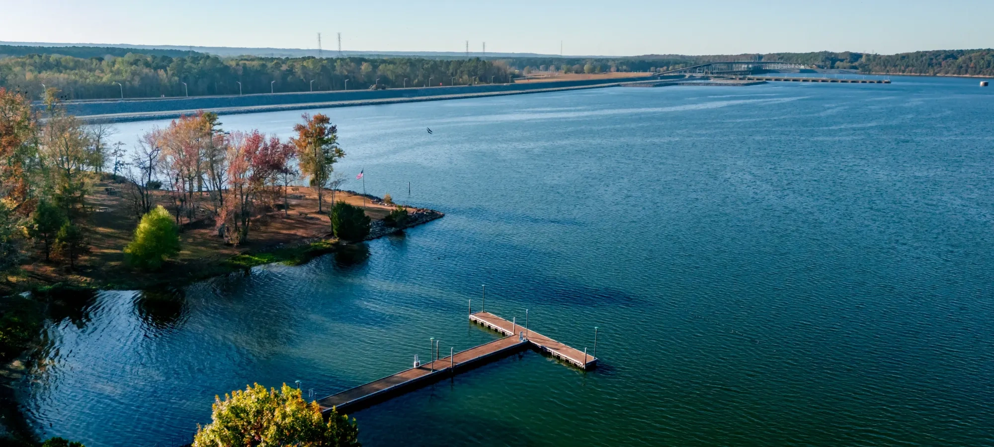 Lake with dock, surrounded by autumn trees under clear blue sky.