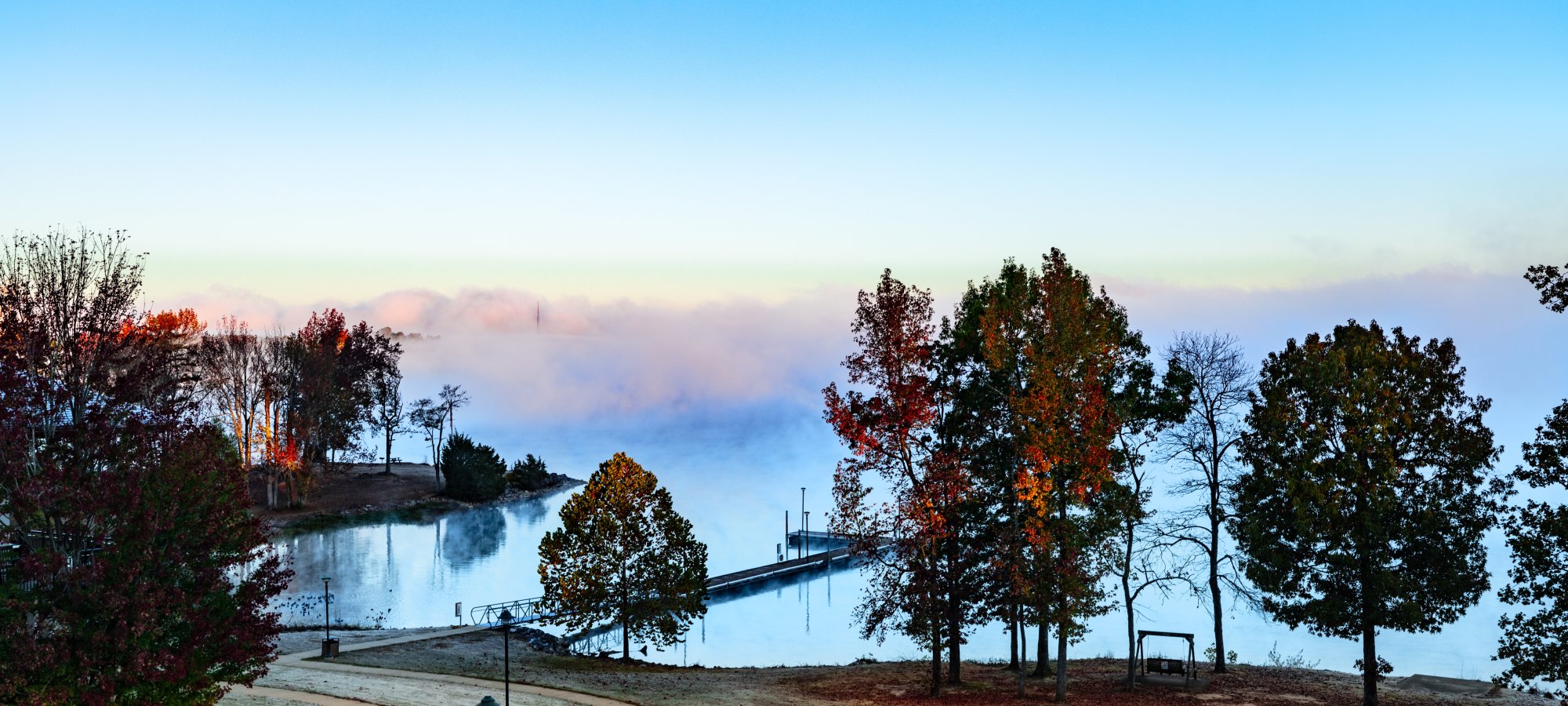 Serene lakeside morning scene with mist rising, vibrant autumn trees, and a dock stretching into calm water. A winding path adds a peaceful tone.