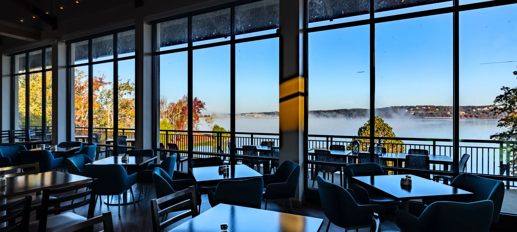 Restaurant interior with large windows overlooking a misty lake and autumn trees.