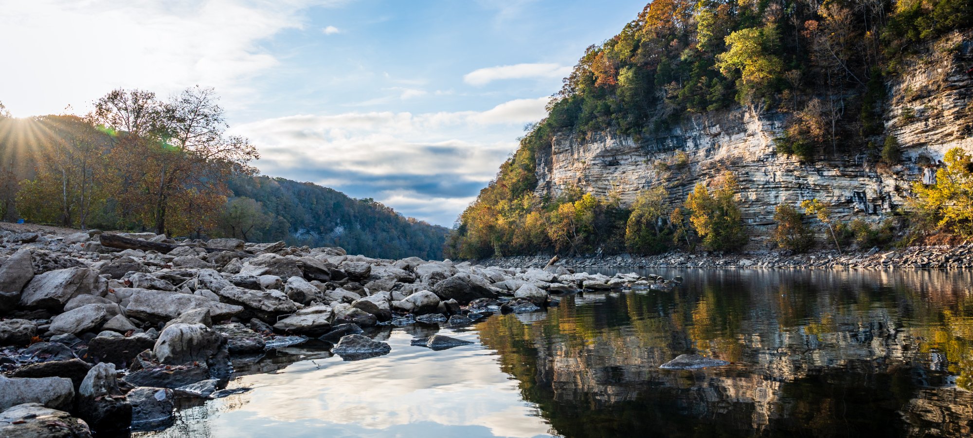 A tranquil river reflects the blue sky and surrounding cliffs, with scattered rocks lining the shore.