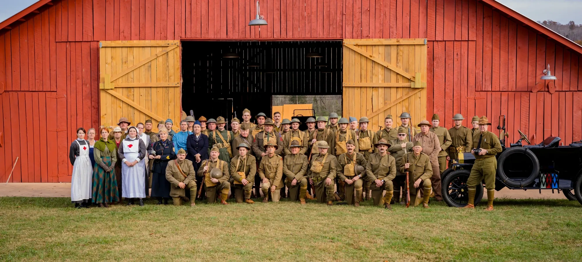 Group photo at a vintage reenactment with people in historical military uniforms and dresses in front of a large red barn, conveying nostalgia.