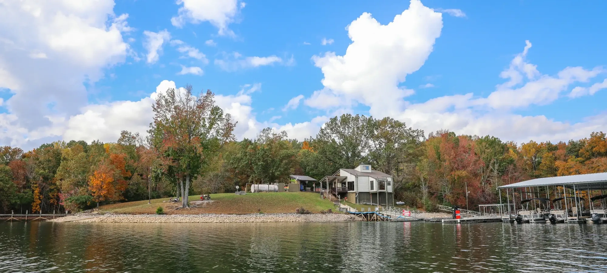 Lakefront house with autumn trees and a dock, under a blue sky with clouds.