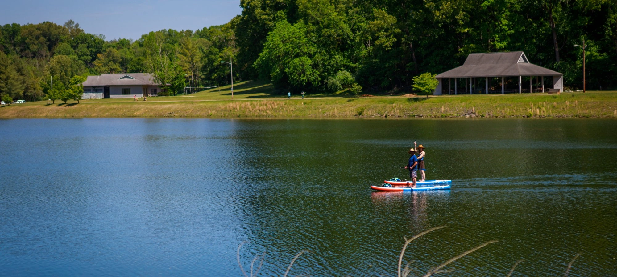 A serene lake scene with two people standing on a blue and red paddleboard. Trees and two buildings are visible in the background under a clear blue sky.