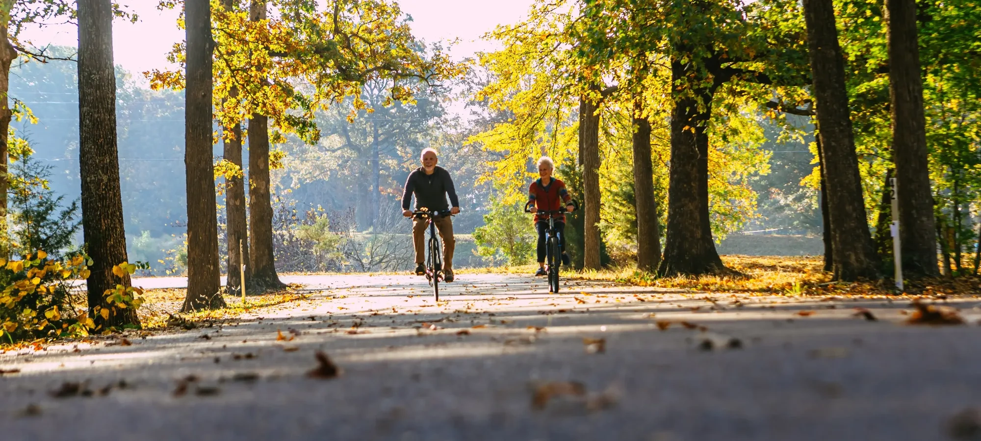 A couple bikes down a wide trail, surrounded by trees, smiling. 
