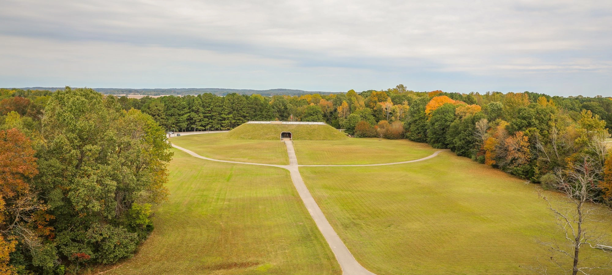 Green field with a path leading to a large grass mound, surrounded by trees with autumn foliage.