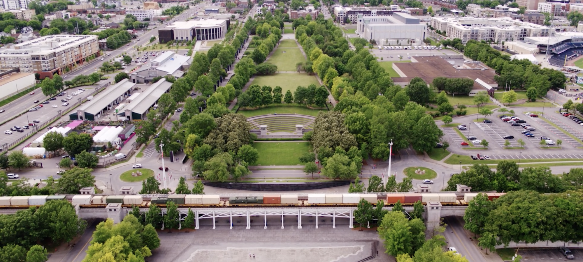 A broad aerial shot shows the entire length of the park, including the amphitheater, long central lawn, Market House buildings to the left, and parking areas to the right, with a freight train passing along the park’s southern edge.