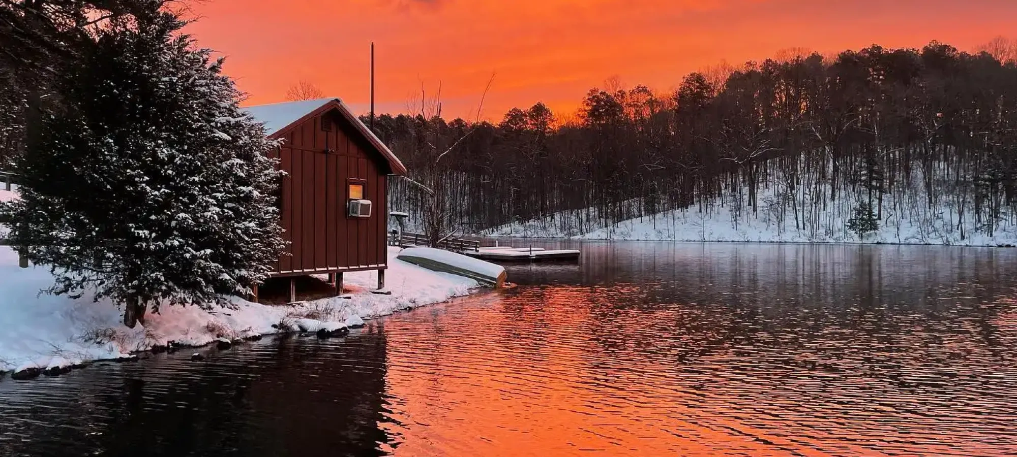 A snowy lakeside cabin with a vibrant, fiery orange sky reflecting in the calm water. Snow-covered trees line the horizon, creating a serene winter scene.