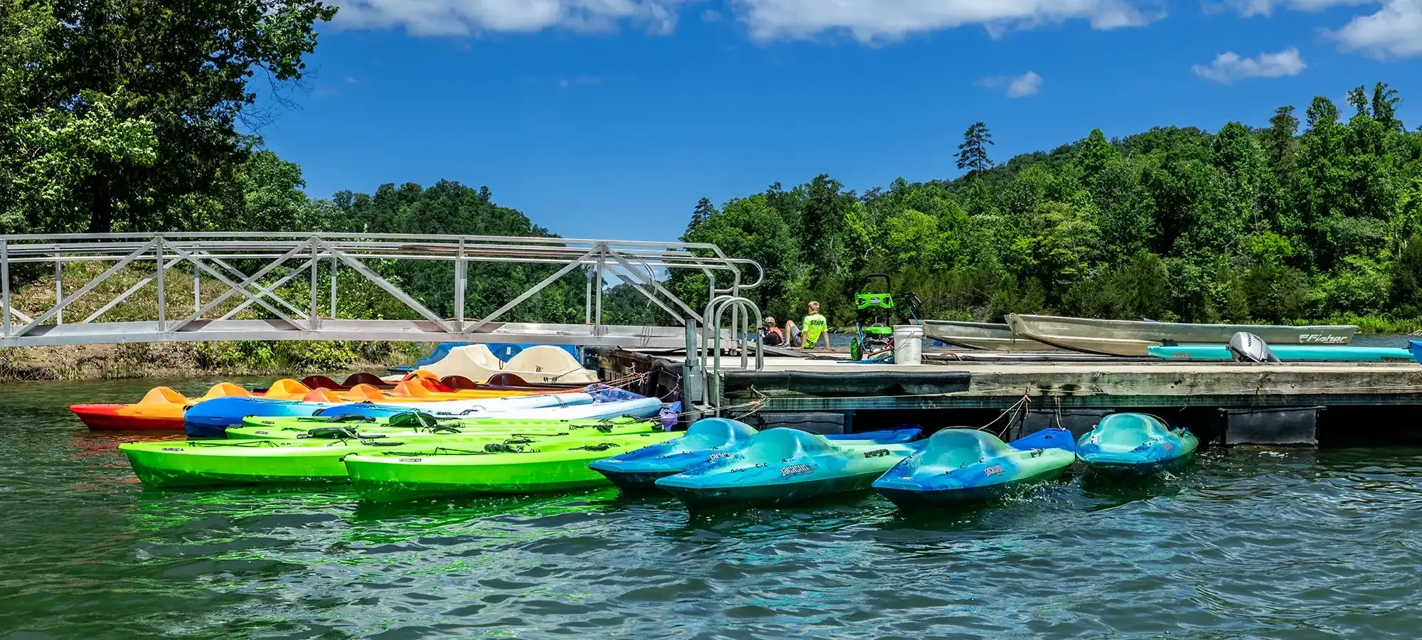 Colorful kayaks and paddle boats are docked on a calm lake under a bright blue sky. The green forest backdrop adds a serene and inviting feel.
