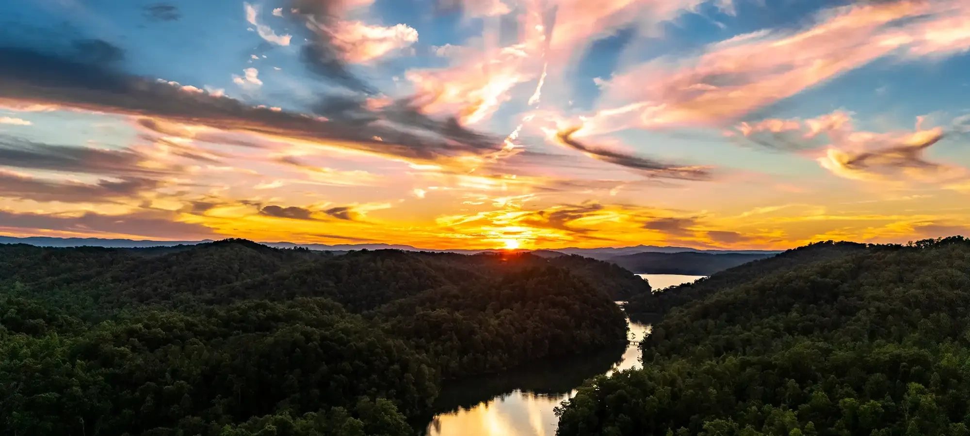 A vibrant sunset over a serene river winding through dense, green hills. Dramatic clouds streak the sky in pink, orange, and blue, creating a tranquil scene.