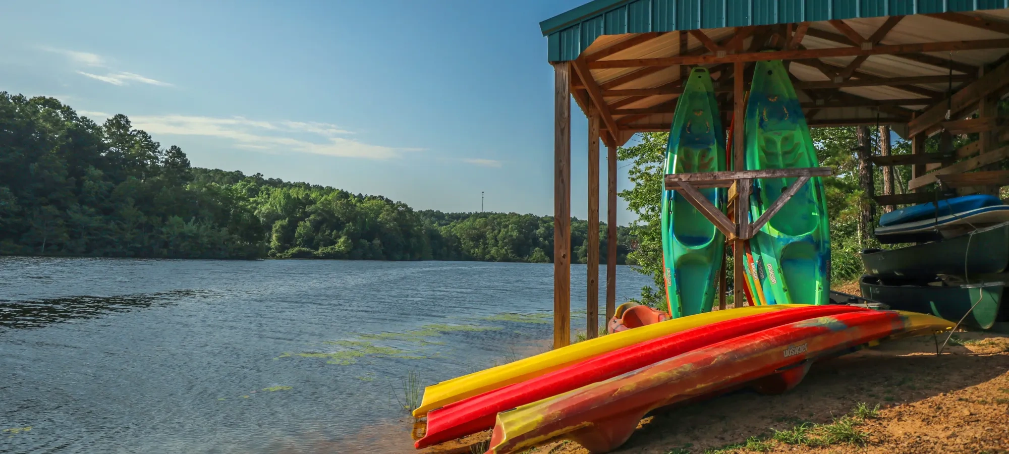 Kayaks and a canoe rest under a wooden shelter by a tranquil lake, surrounded by lush trees. The scene is peaceful and sunlit under a clear blue sky.