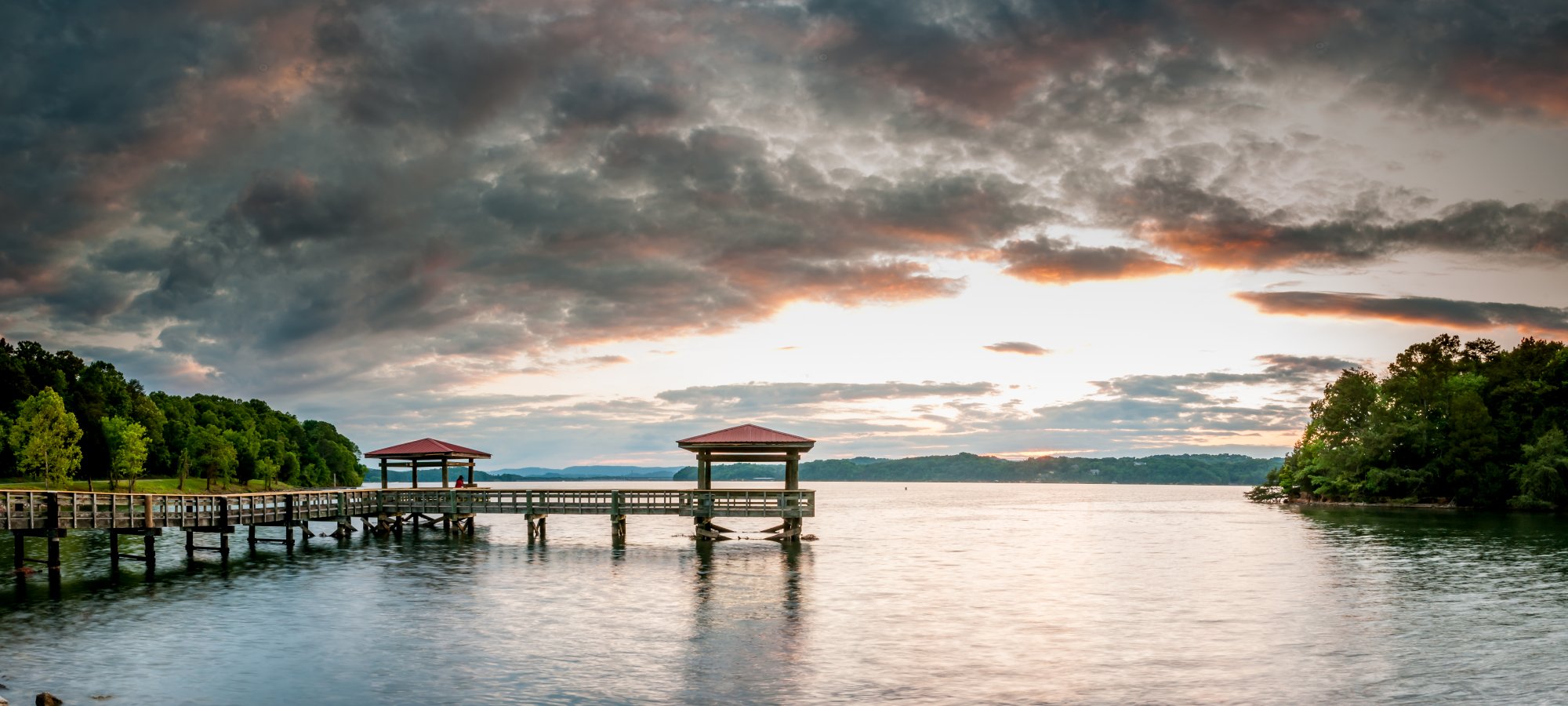 Sunset over a calm lake with wooden piers extending into the water. Dramatic clouds fill the sky, surrounded by lush green trees, creating a serene atmosphere.