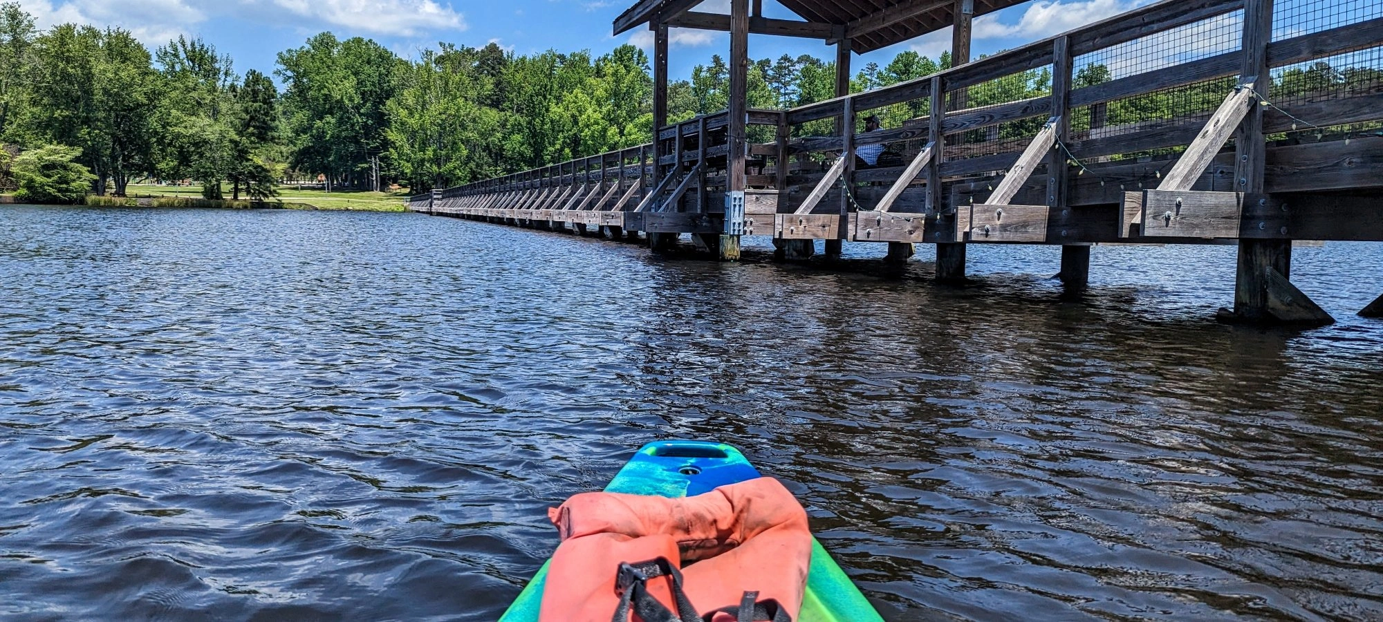 Green kayak with an orange life jacket on a lake near a wooden bridge under a blue sky.