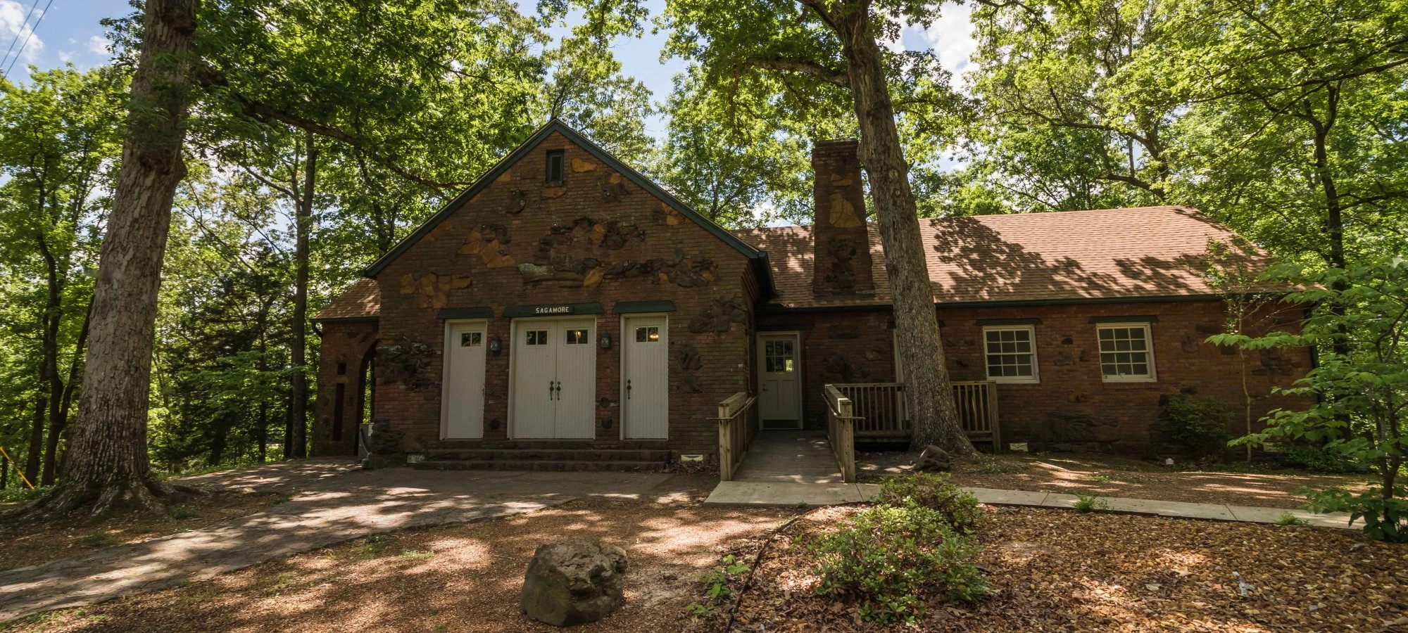 Rustic brick lodge in a forest setting, surrounded by tall trees and sunlight.