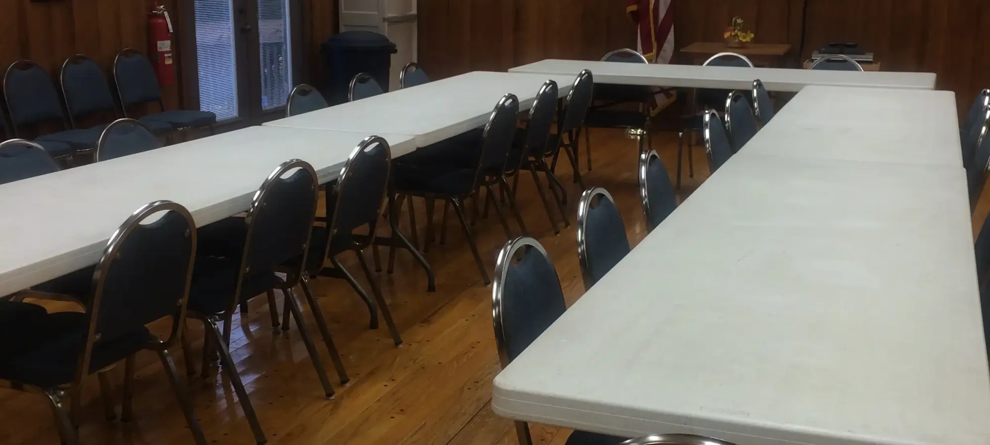 Meeting room with tables, chairs, wooden floor, and American flag.